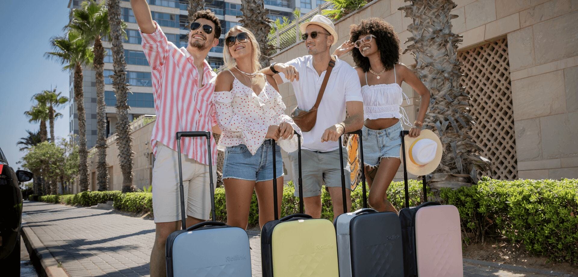 Four travelers with rolling suitcases smiling and taking a selfie outside a sunny palm-lined hotel
