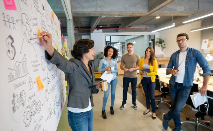 An office full of smiling people. One woman is pointing at a white board full of sticky notes and scribbles, while a team of men and women are watching her, listening to her.