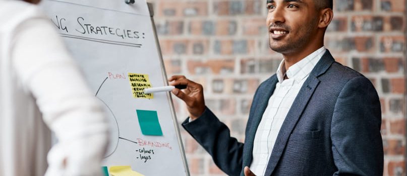 A confident man in a suit is standing in front of a white board, pointing at it while talking.