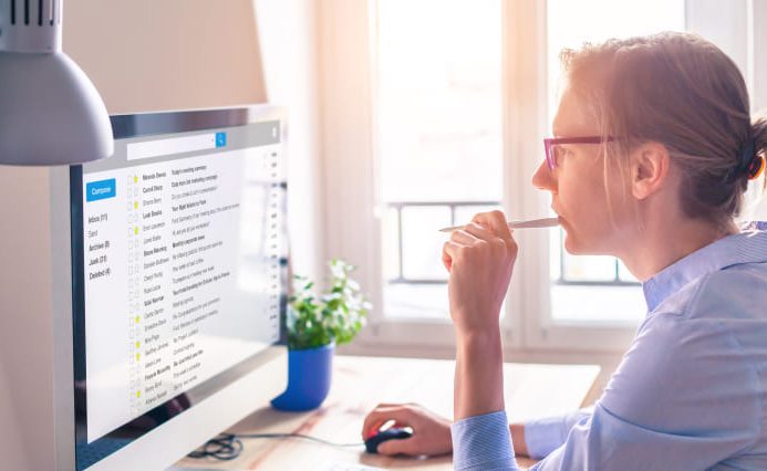 A woman is sitting at a desk, looking at a computer screen filled with data. Deep in thoughts, she put her pen to her mouth.