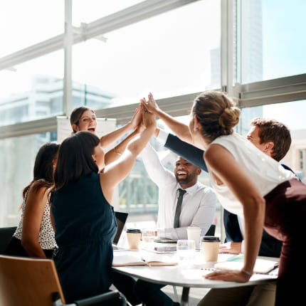 A professionally dressed team of people is high-fiving over a desk.