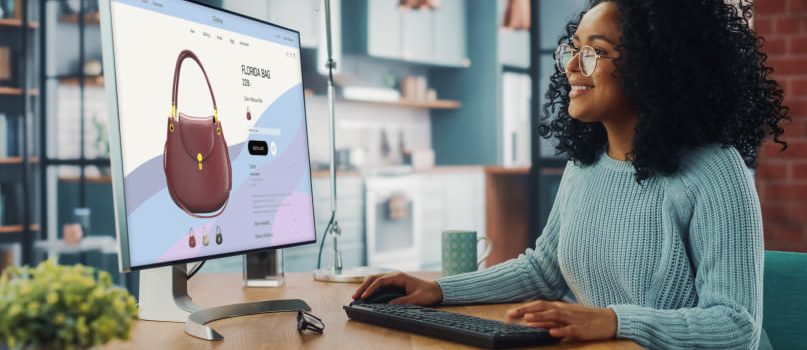 A woman is sitting at her desk, smiling, and designing an ecommerce page on her computer. There is a brown handbag on the screen.