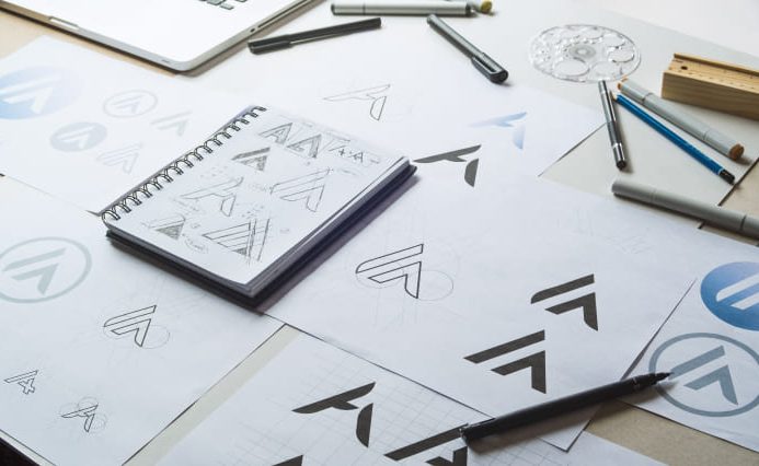 A notebook and a bunch of white pieces of paper are scattered across a desk, featuring the notes and prints by a professional logo design company.