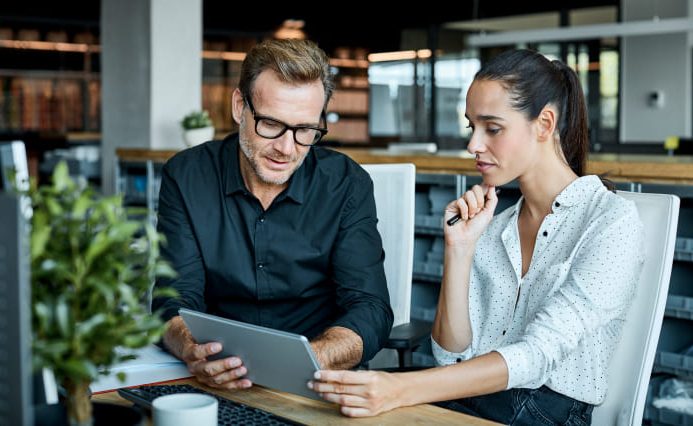 A professional-looking man and woman are sitting together at a desk, looking at a tablet and having a discussion.