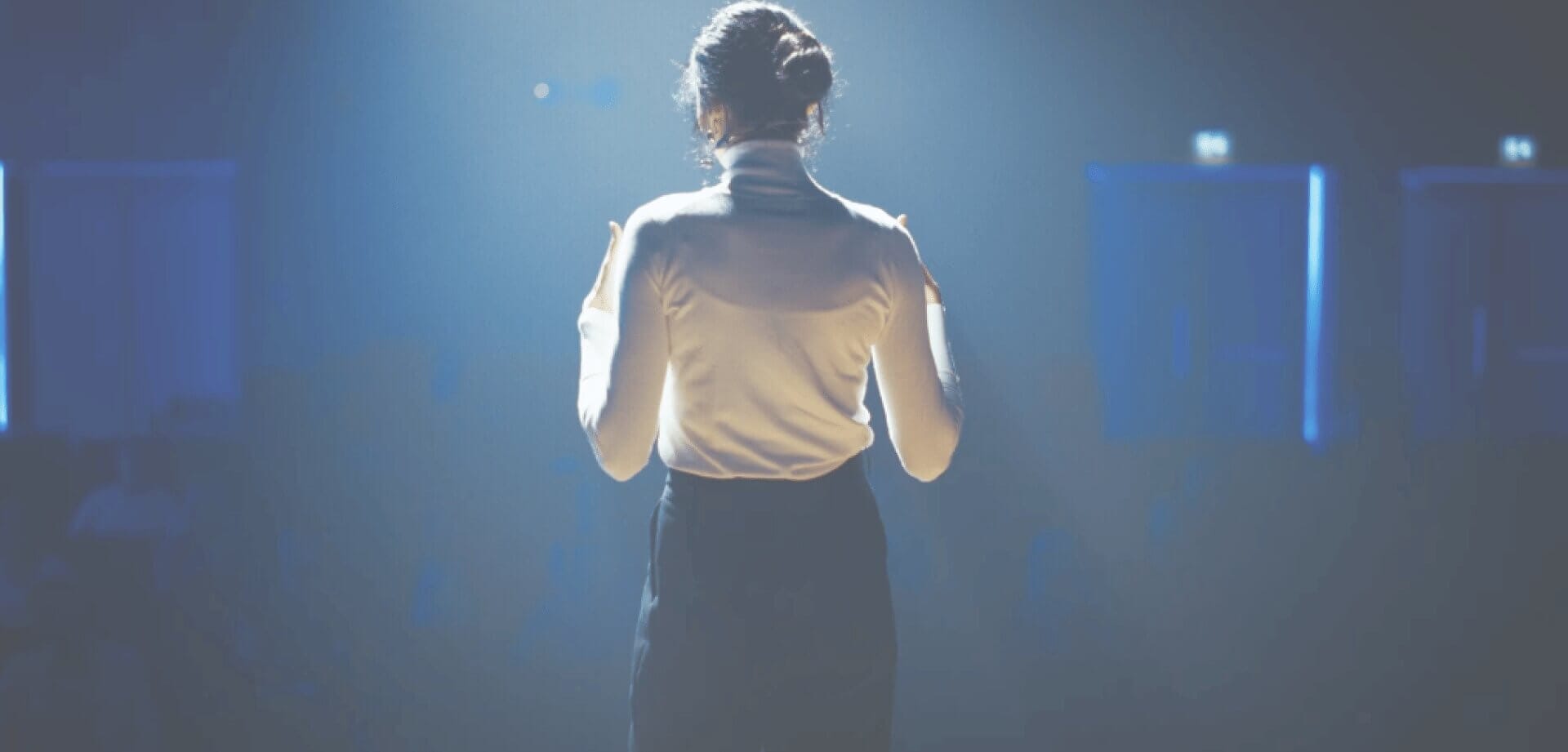 Professional speaker standing on stage under bright spotlight, addressing an audience at a conference or corporate event in a large auditorium.