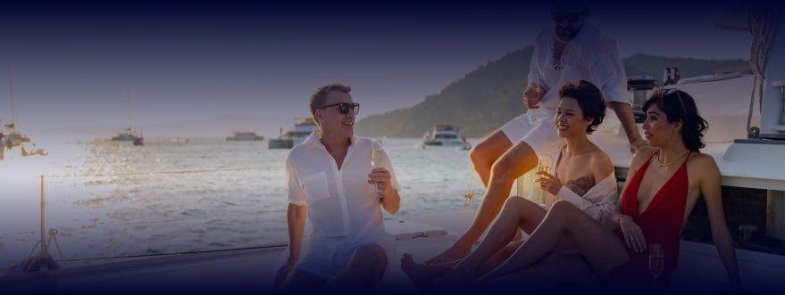 Group of friends relaxing on a yacht at sunset, holding drinks with boats and hills in the background