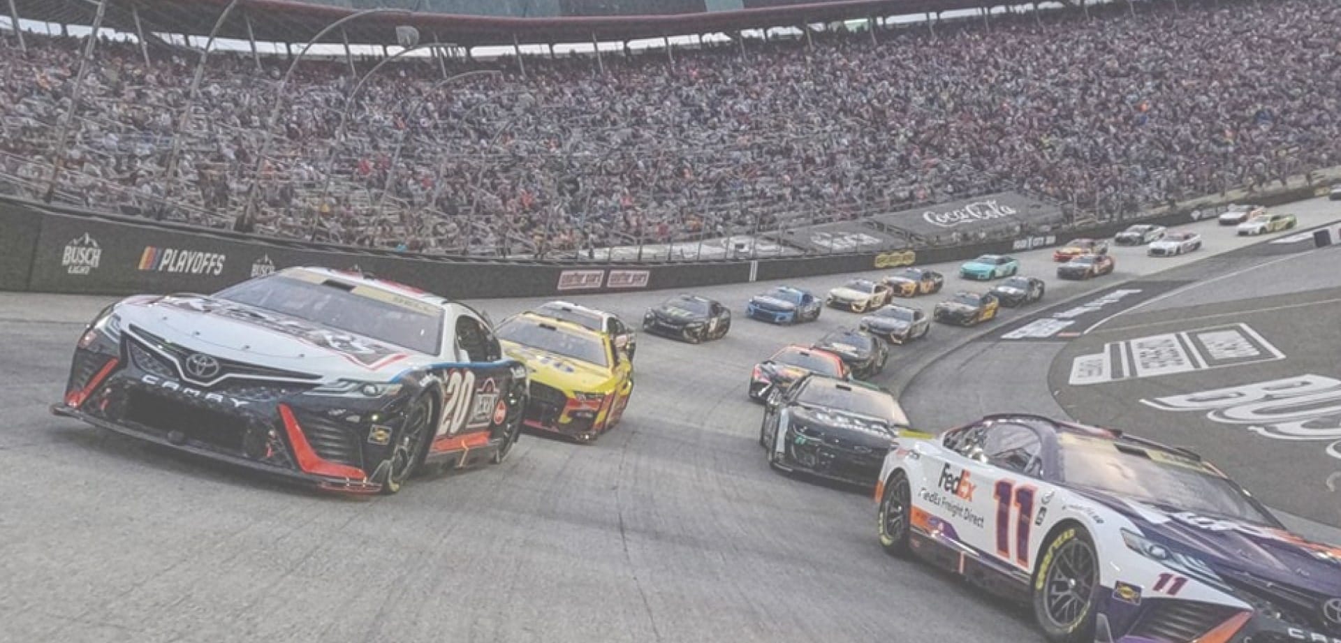 Stock cars racing in a pack on a banked oval track, with a full grandstand in the background