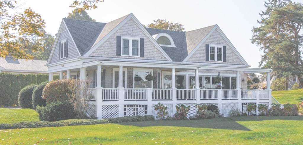 Cape Cod style home with a wraparound porch, shingle siding and manicured lawn on a bright autumn day.