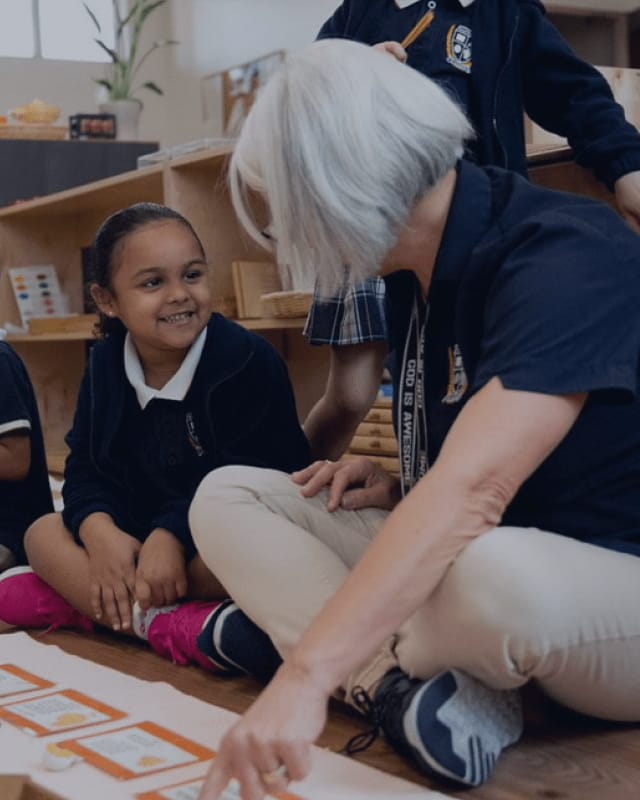 ISP-tall-background A teacher sitting on a classroom floor working through a learning activity with a smiling young student in school uniform.