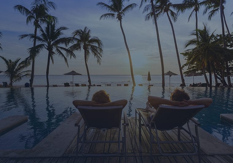 Two people relax in lounge chairs beside an infinity pool overlooking the ocean at sunset