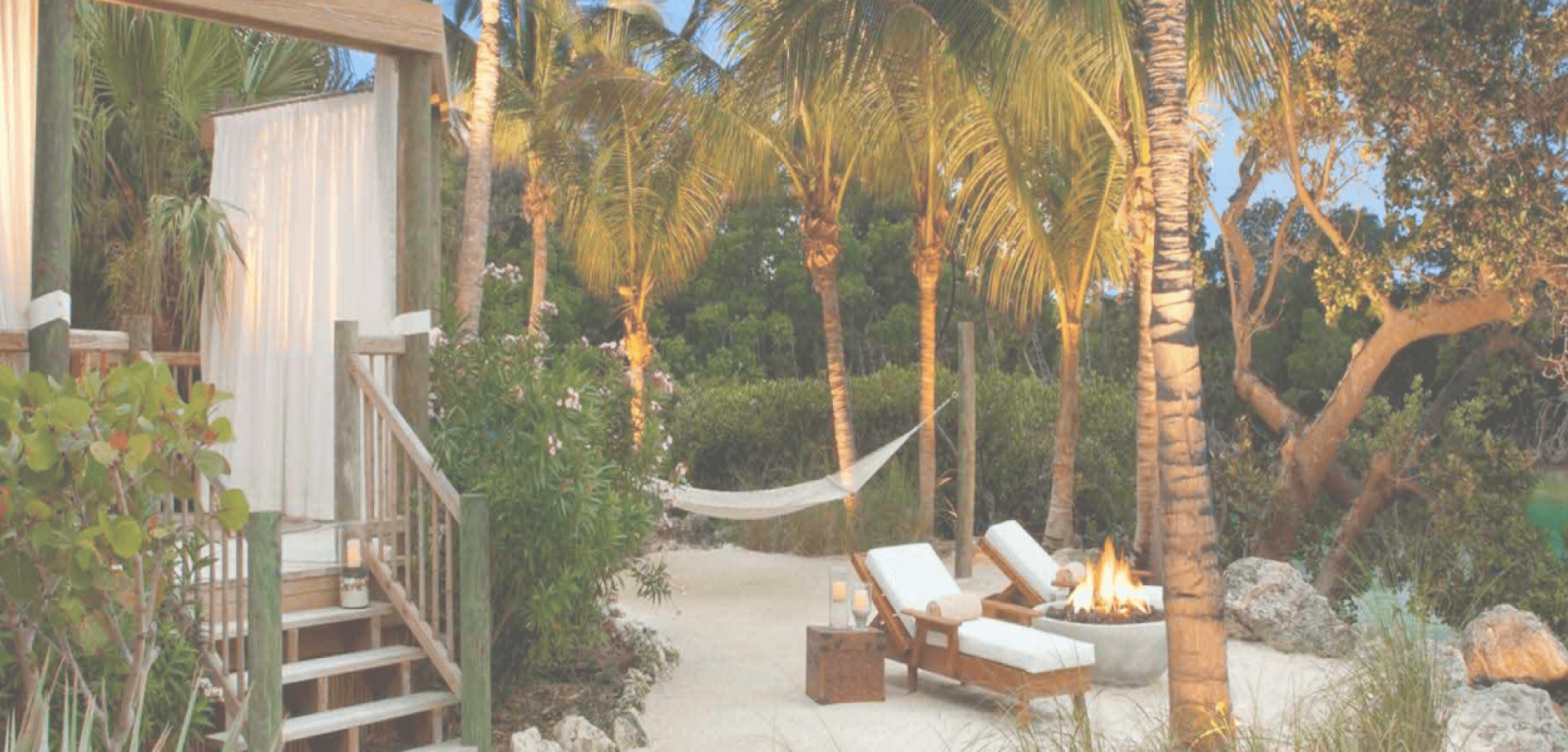 Tropical outdoor lounge area with palm trees, a hammock, cushioned chairs, a fire pit, and a cabana-style structure