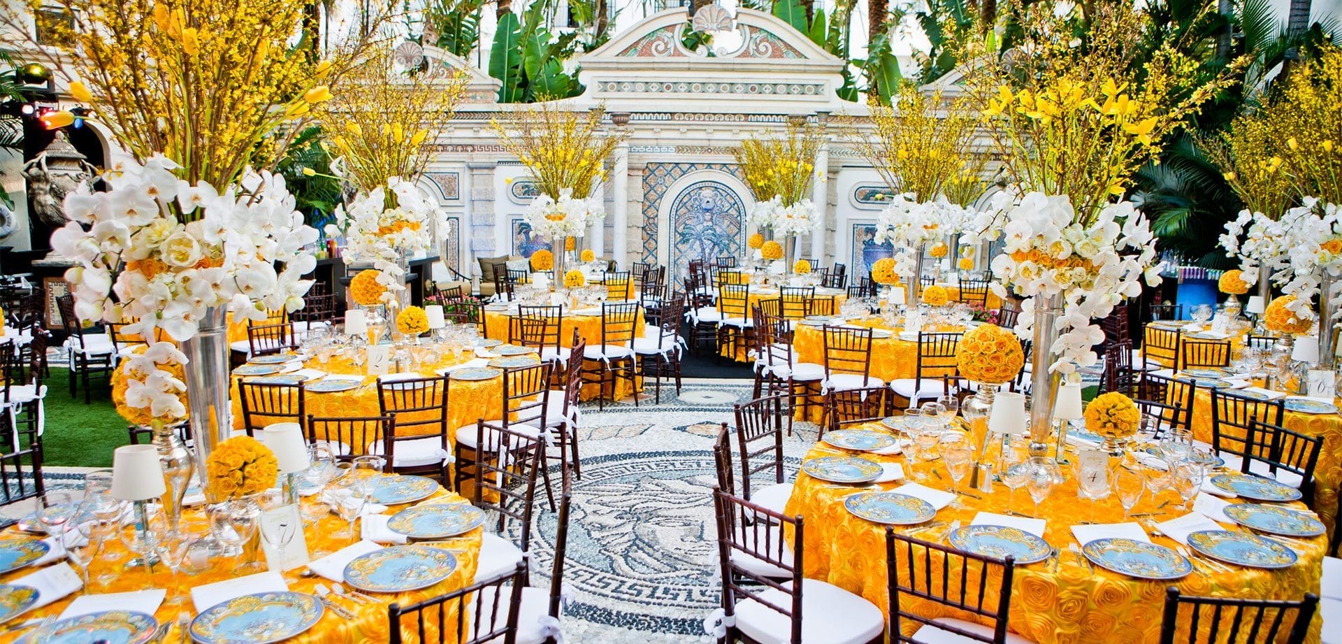 Elegant outdoor dining setup with round tables, bright yellow table linens, floral centerpieces, patterned plates, and ornate architectural details