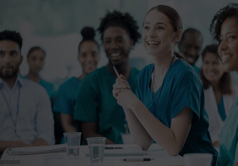 Smiling healthcare workers seated together in a group discussion, with one woman in scrubs holding a pen and speaking.