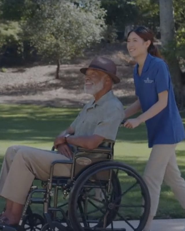 Caregiver pushing an elderly man in a wheelchair along a paved path in a sunny park.