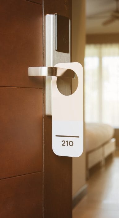 Hotel room door with a hanging door tag marked 210, with a bed visible in the background.