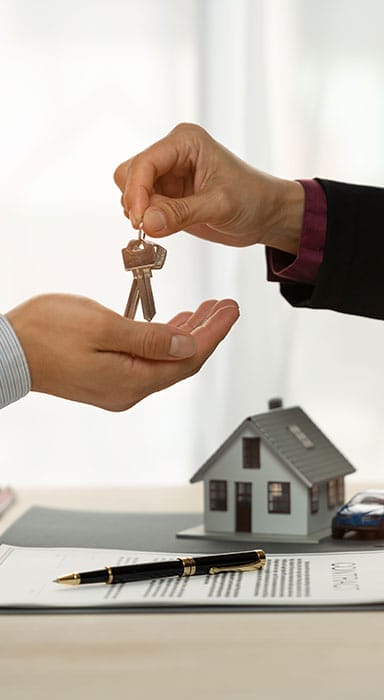 Real estate agent handing house keys to a client above a property contract, with a model home and pen on the desk.