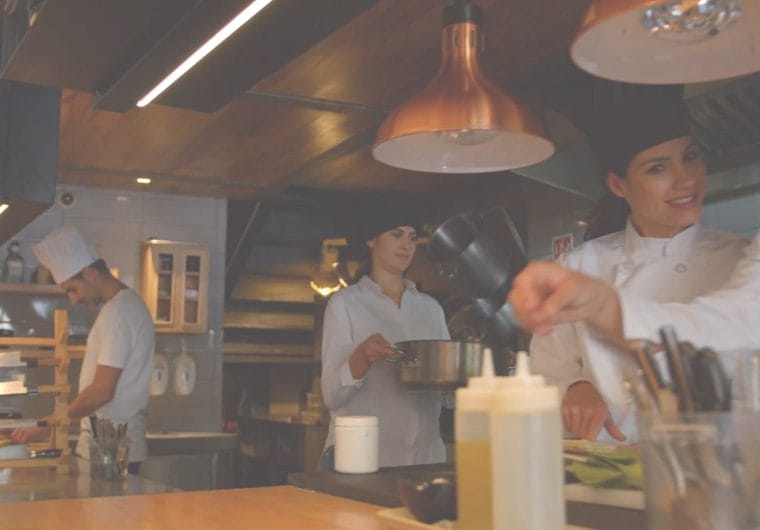 education-website-design-culinary-academy-of-las-vegas-background-1 Culinary students and chefs working together in a professional kitchen with cookware, ingredients, and warm lighting.