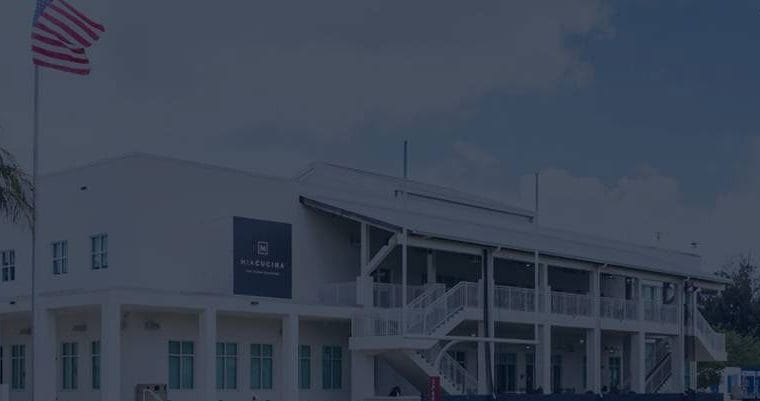 education-website-design-miami-country-day-school-wide Wide exterior view of a Miami Country Day School campus building with palm trees, an American flag, and a sports field light in the background.
