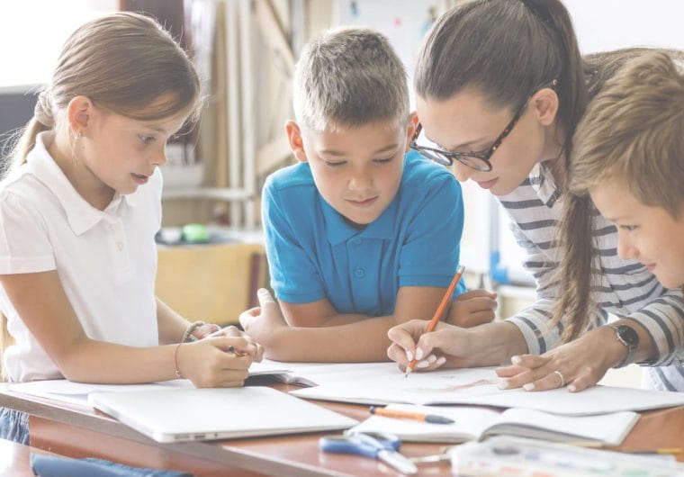 education-website-design-savvas-background-image A teacher and three students gathered around a desk working together on a writing activity in a bright classroom.