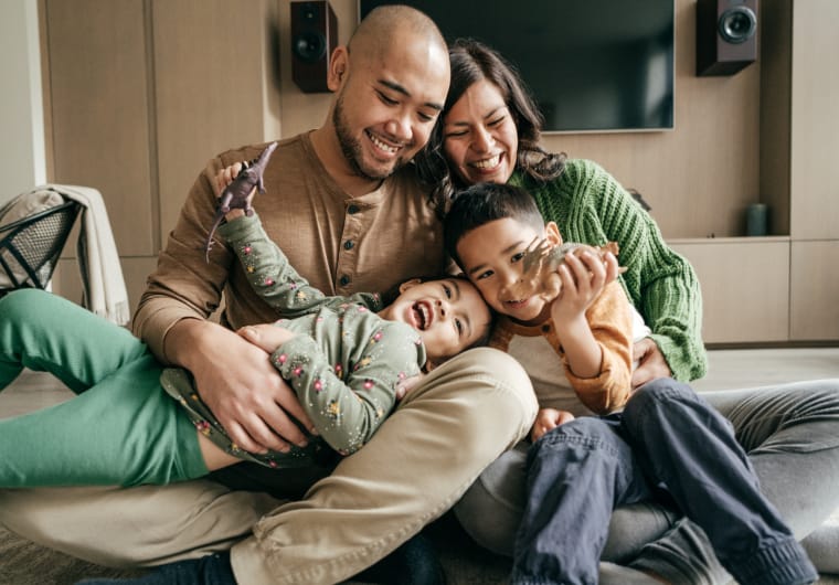A smiling family of four sits on the floor in a cozy living room, hugging close together while two children play with toy dinosaurs