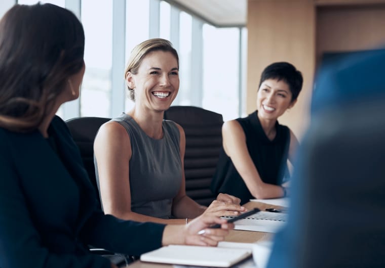Three women sitting around a conference table in a bright office, smiling and engaged in conversation during a meeting