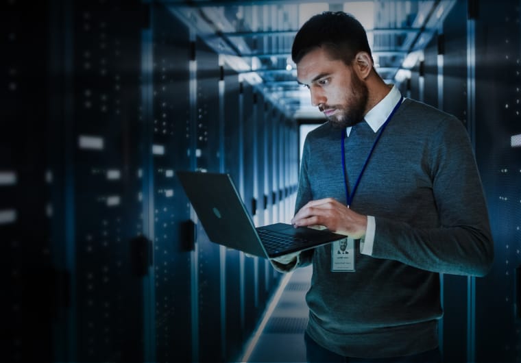 A badged IT professional standing in a server room corridor, working on a laptop amid rows of illuminated data center equipment