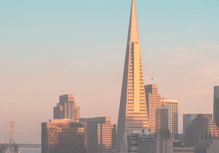 The San Francisco skyline at golden hour, featuring the Transamerica Pyramid tower prominently against a soft peach and blue sky, with the Bay Bridge visible in the background