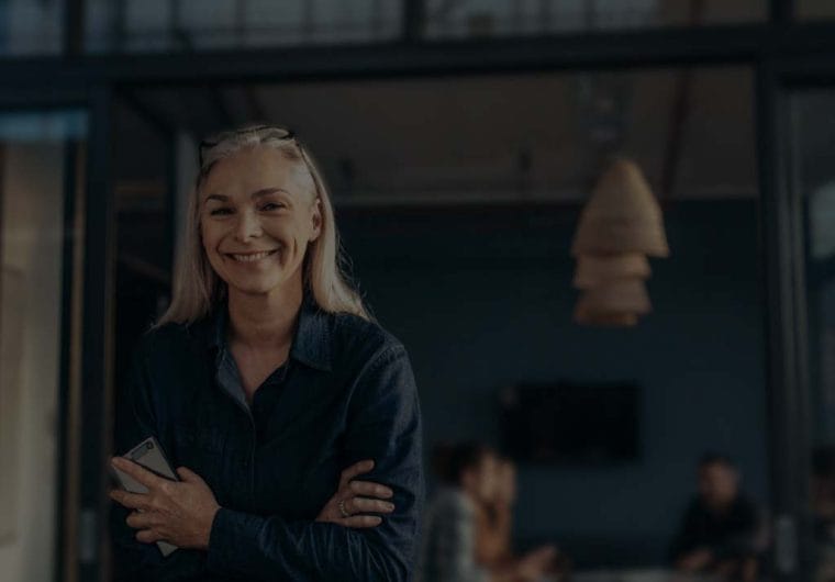 A confident businesswoman with silver-blonde hair standing in a modern workspace, arms crossed, holding a smartphone and smiling