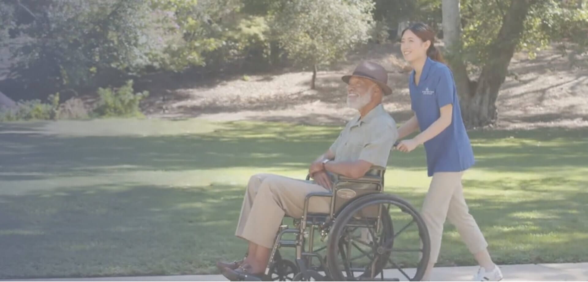 Senior caregiver assisting an elderly man in a wheelchair during an outdoor walk through a sunny park, highlighting compassionate mobility support and personalized senior care services.