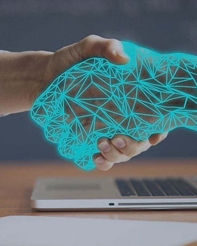 Boston Materials 33 Human hand reaching toward a glowing digital wireframe hand above a laptop, representing AI technology and human collaboration