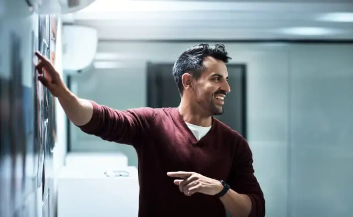 Man in a maroon shirt smiling while writing on a glass board in a bright office