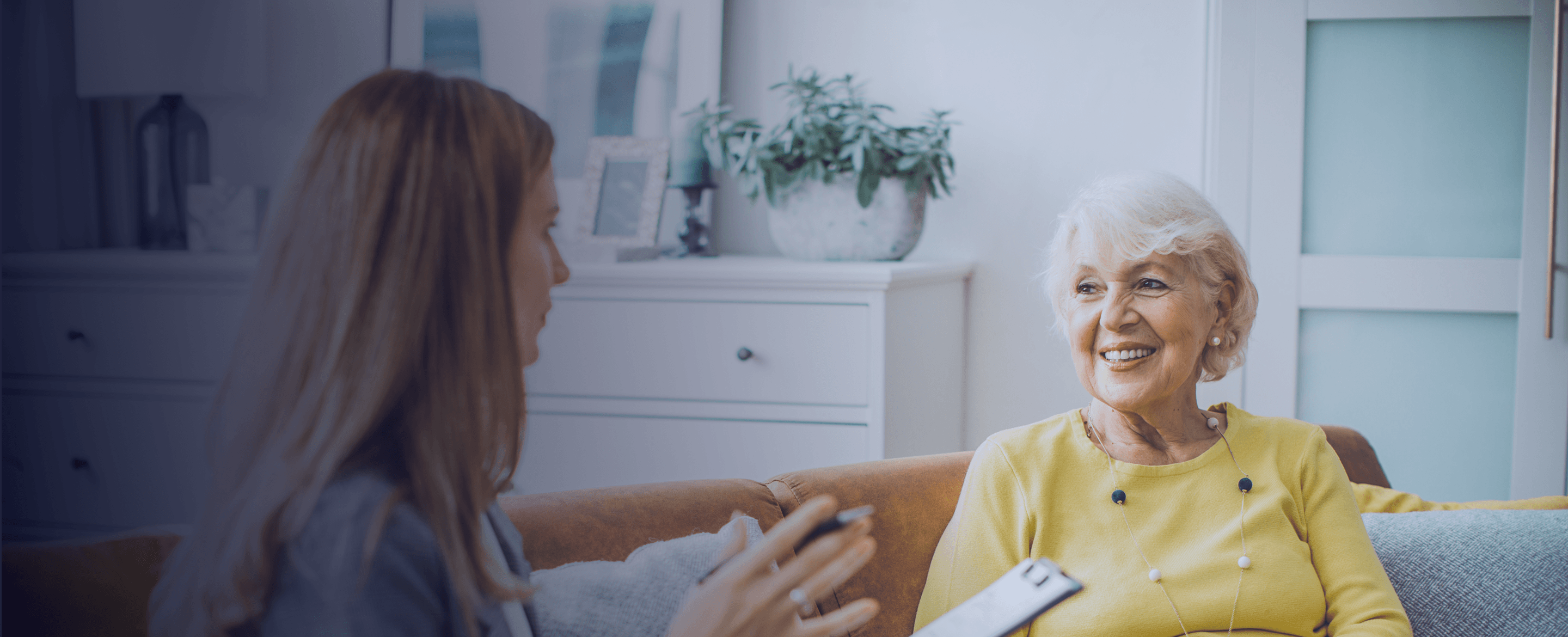 Older woman smiling while speaking with a younger woman holding a clipboard in a living room.