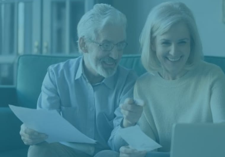 Older couple sitting together on a couch, smiling at a laptop while holding papers