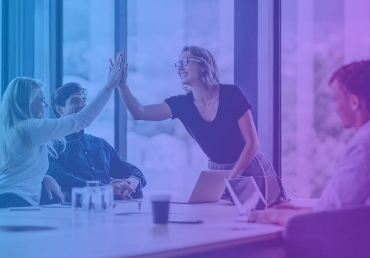Two colleagues high-fiving across a conference table in a bright modern office, with two other team members smiling nearby