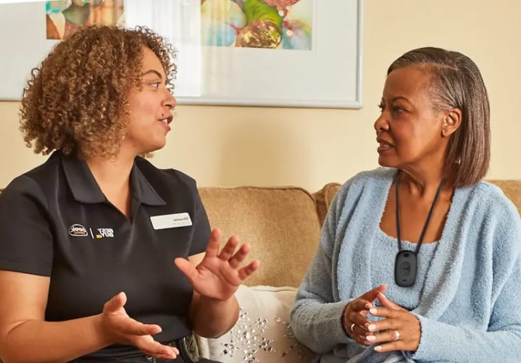A care worker in a black uniform talks with an older woman seated on a couch
