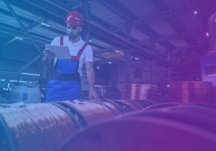 A factory worker wearing a hard hat and overalls checks a tablet beside large spools of wire in an industrial warehouse
