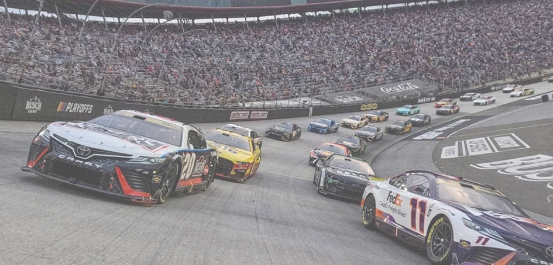 Stock cars racing closely together on an oval track in front of a packed grandstand