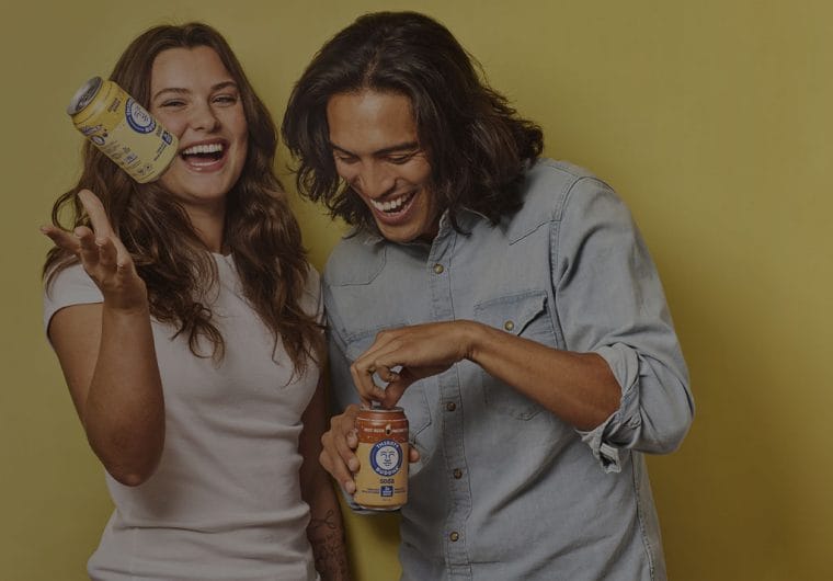 Two laughing people holding Thirsty Buddha soda cans against a warm yellow background, with one tossing a can in the air