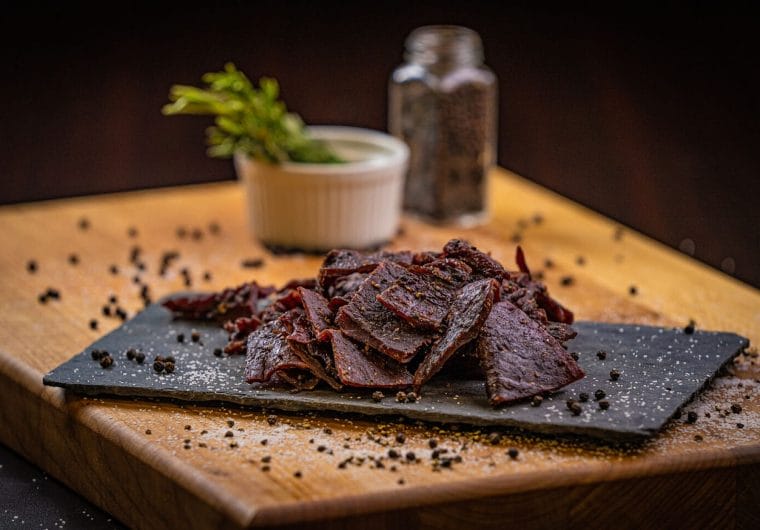 Sliced beef jerky pieces stacked on a slate board over a wooden surface, scattered with black peppercorns and seasoning, with a small ramekin and spice jar in the background