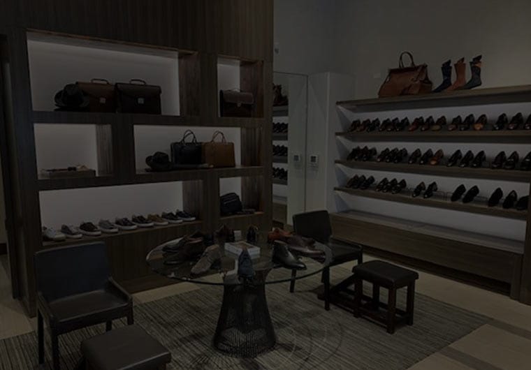 Dimly lit interior of a luxury retail shoe and accessories boutique with dark wood shelving displaying leather dress shoes, loafers and handbags across two walls