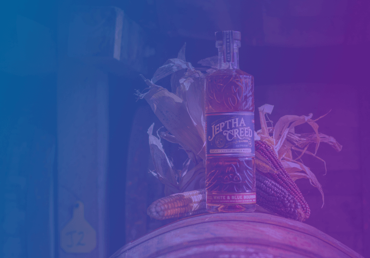 A bottle of Jeptha Creed bourbon stands on a wooden barrel surrounded by ears of corn