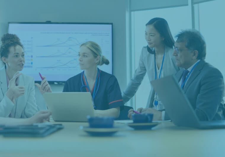 Four healthcare professionals including a nurse, a physician and two executives collaborating around a laptop in a meeting room with data charts visible on a screen behind them, overlaid with a blue and purple gradient