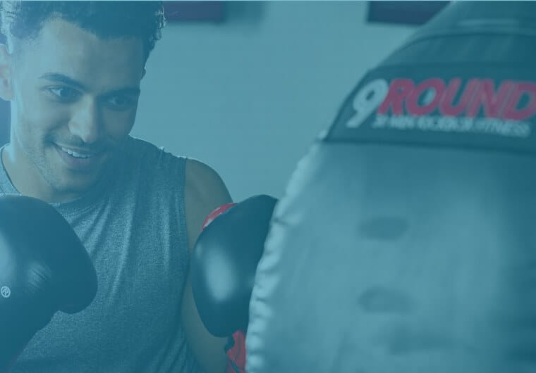 A smiling man wearing boxing gloves trains with a punching bag in a softly lit gym