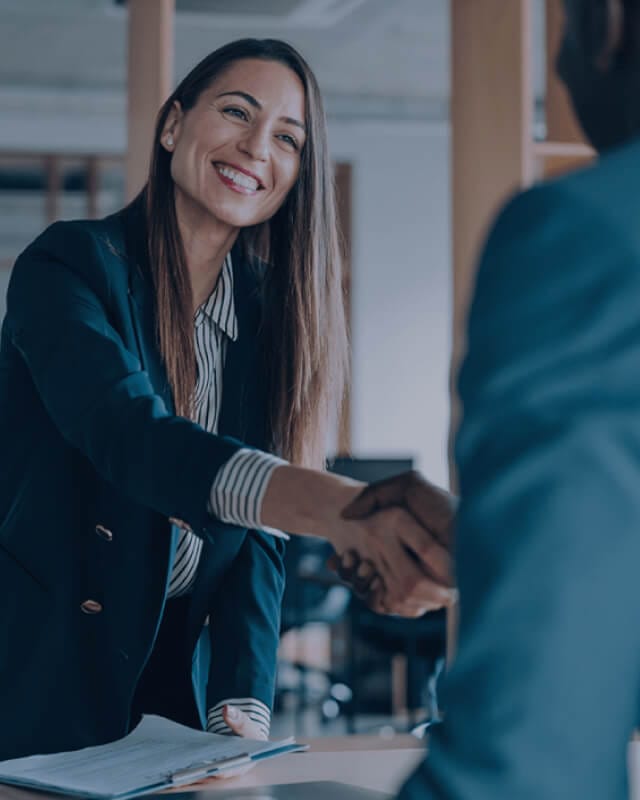 smiling businesswoman in a navy blazer shaking hands with a client across a desk in a professional office setting