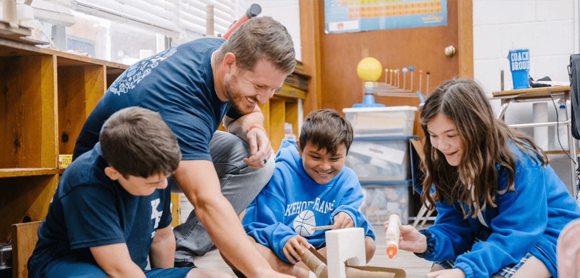 A teacher crouches beside three students as they work together on a hands-on classroom project using cardboard, glue, and craft materials