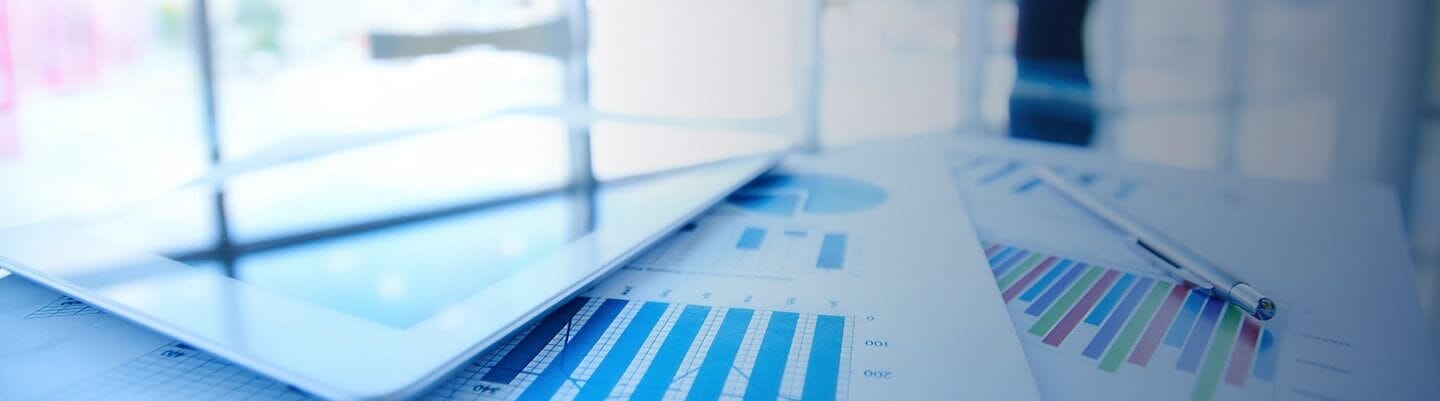 Blurred business analytics background featuring charts, graphs, a tablet device, and financial reports in cool blue tones.