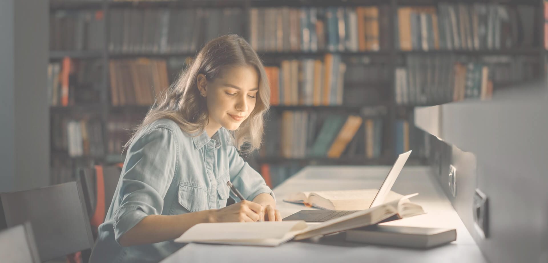 A person writing notes at a library desk with an open laptop and books nearby