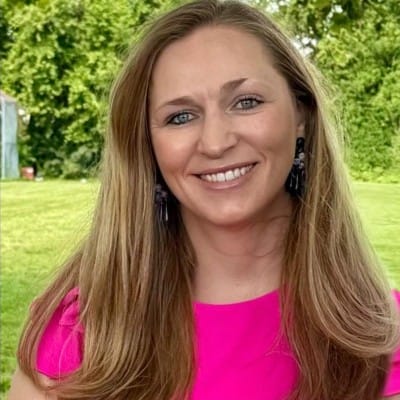 Home Page - NEW 297 Smiling woman with long light-brown hair wearing a bright pink top and black earrings, standing outdoors in front of green trees and a lawn