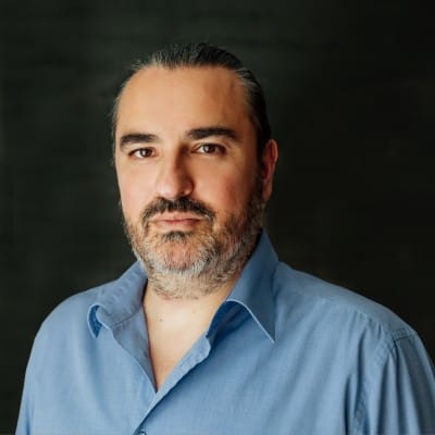 Home Page - NEW 297 Portrait of a man with dark slicked-back hair and a salt-and-pepper beard, wearing a light blue button-down shirt against a dark background