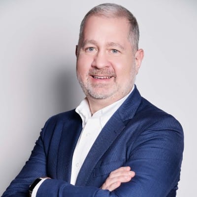 eCommerce Social Media Marketing 59 Portrait of a smiling man with short gray hair and a trimmed beard, wearing a blue blazer over a white collared shirt with his arms crossed against a plain light background